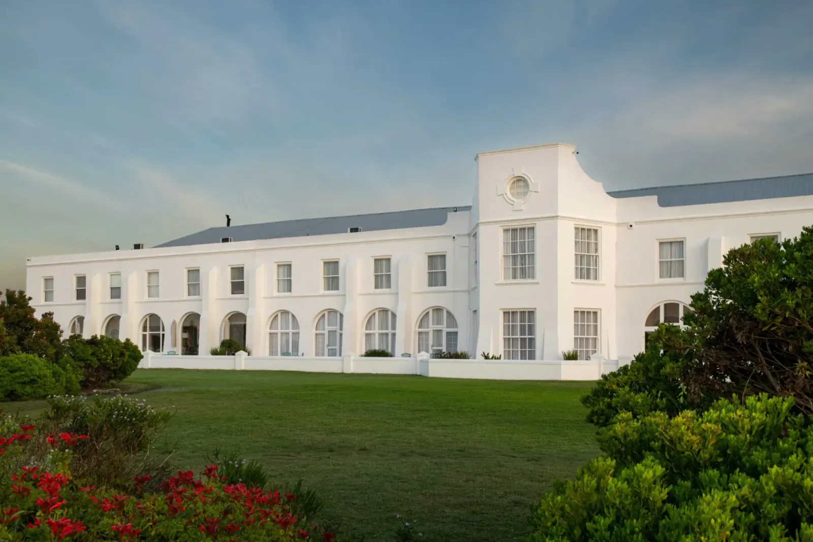 White colonial Marine Hotel in Hermanus, South Africa, with arched windows, clock tower, green lawn, and colorful bushes under cloudy sky.