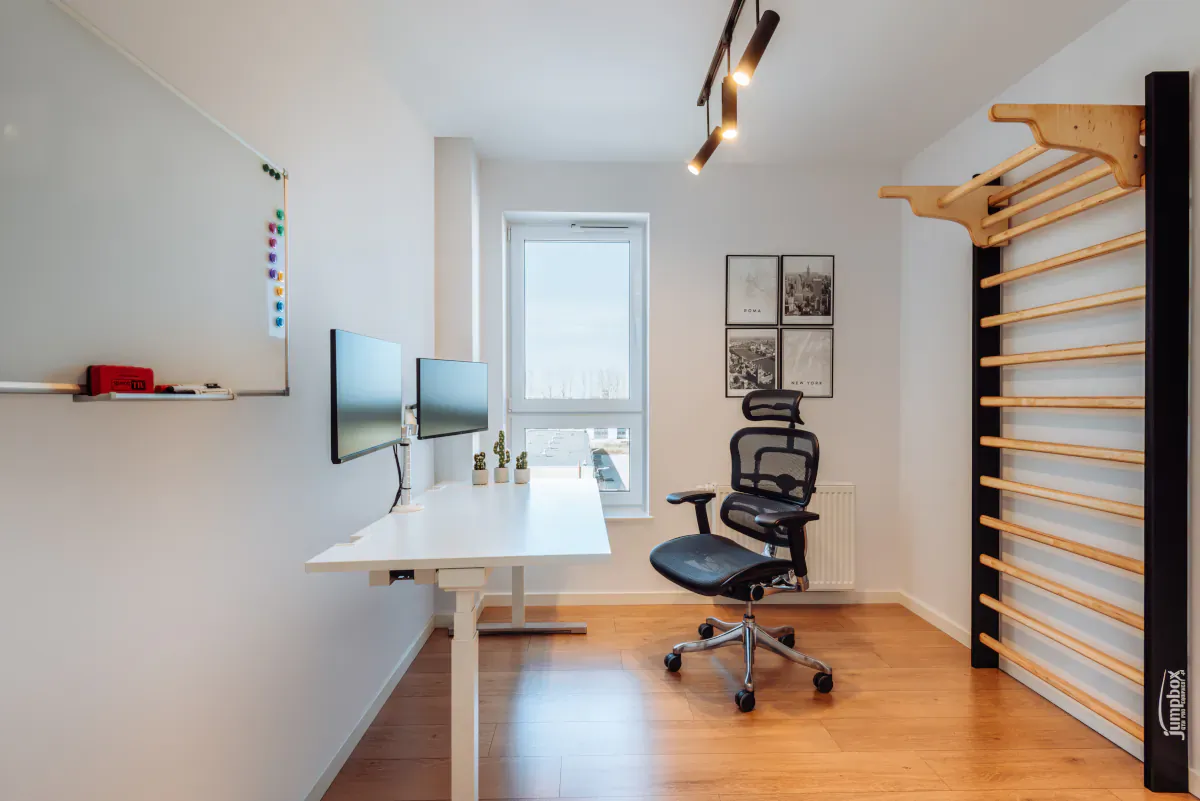 Modern home office with standing desk, dual monitors, ergonomic chair, whiteboard, and wooden wall bars near window.
