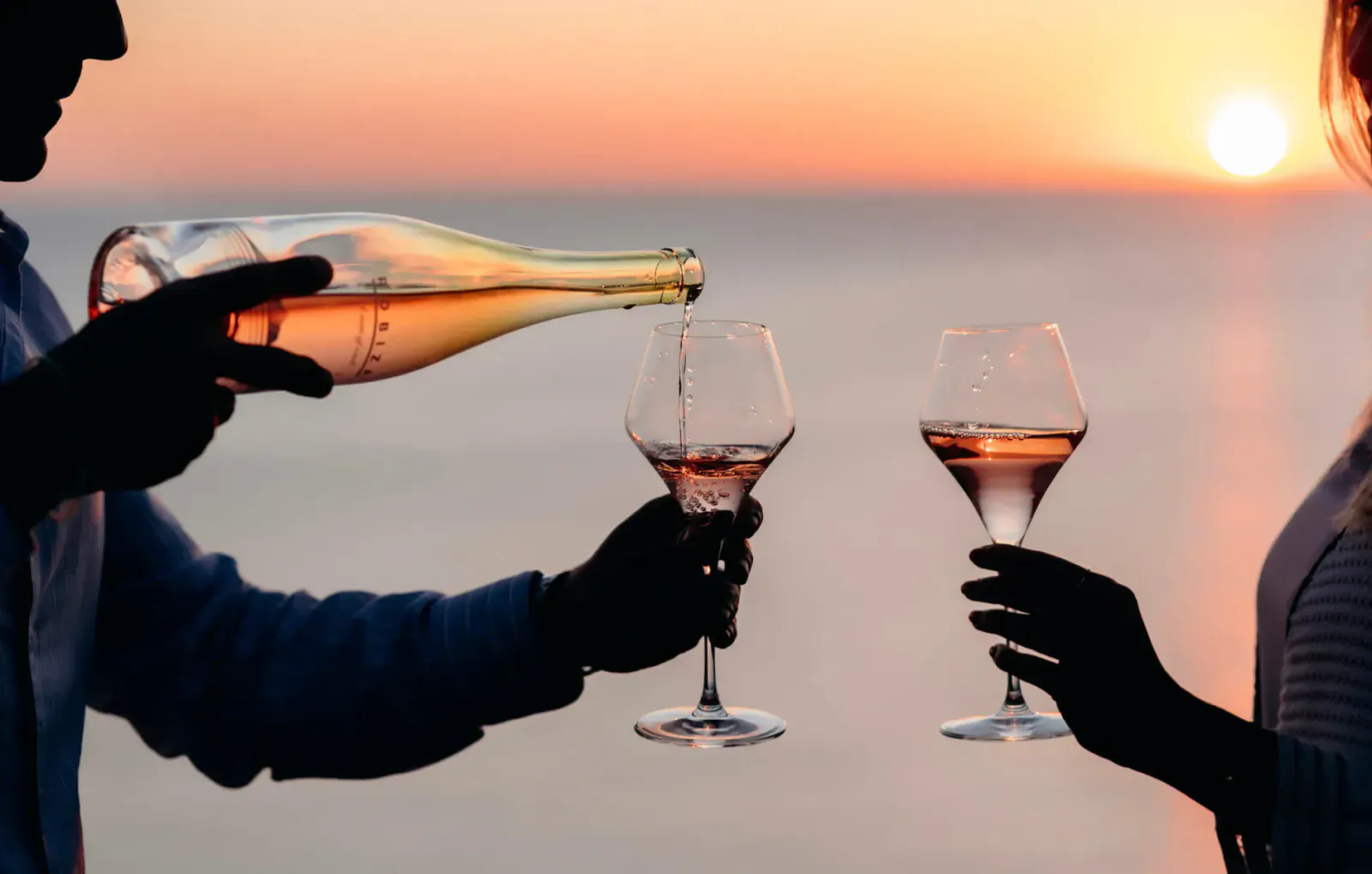 Silhouettes of couple toasting rosé wine at sunset over sea, man pouring from bottle into glasses.