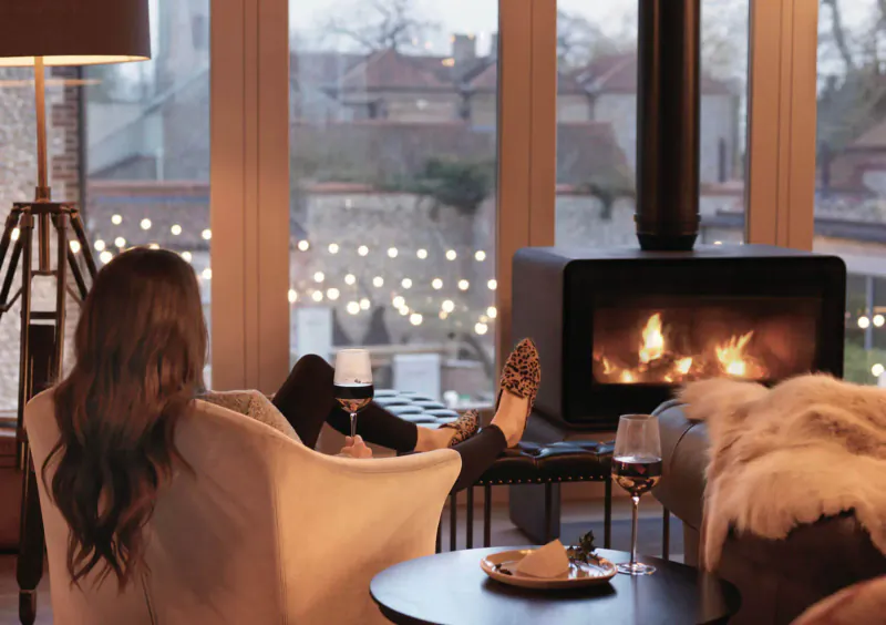 Woman relaxing in cream armchair by fireplace in cozy Harper Norfolk room, feet up, wine glass, winter city view.