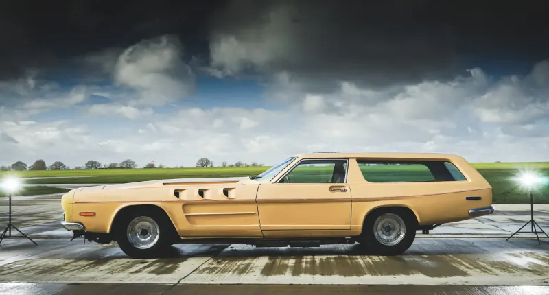Yellow 1970s Ford Falcon XY wagon with V8-style hood scoops on wet pavement, lit by spotlights under cloudy sky