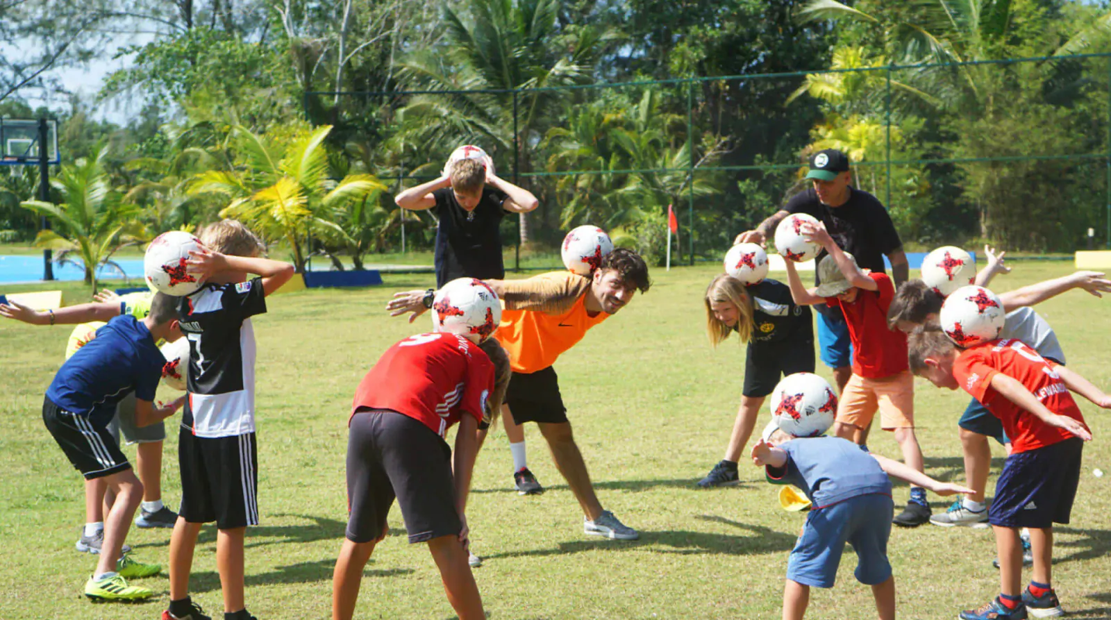 Marcel Gurk, football freestyle expert, leads kids in ball-balancing stretches on grassy court amid palm trees at beach resort.