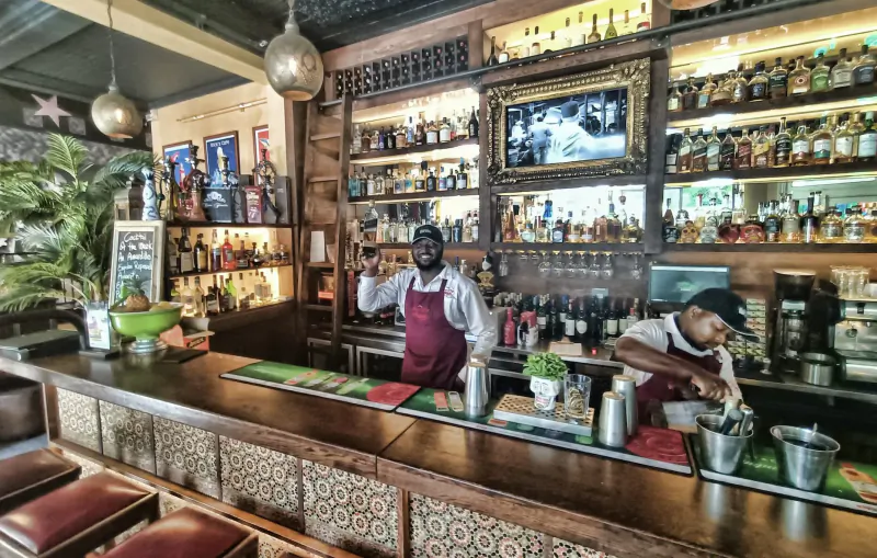 Two bartenders in aprons at a rustic bar with wooden shelves stocked with colorful liquor bottles and pendant lights.