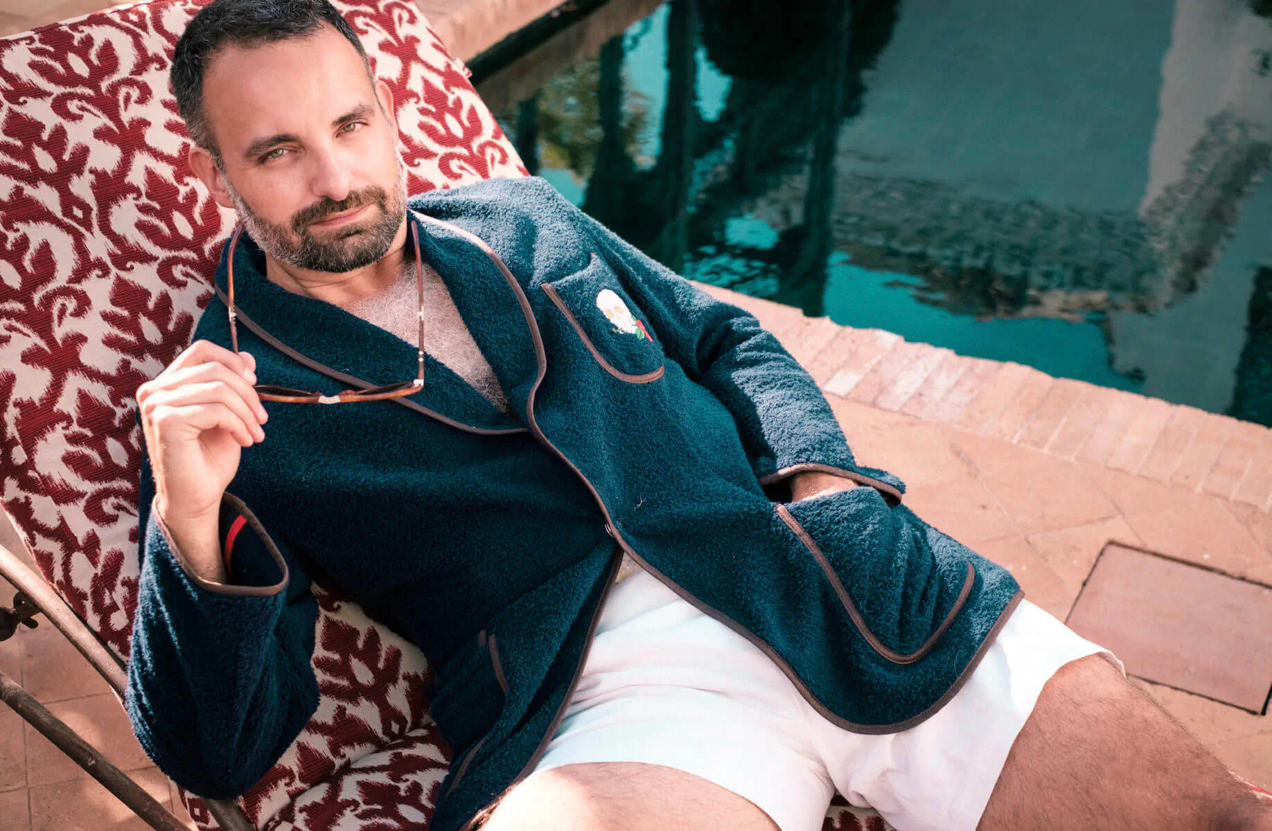 Man in navy Savile Row-style robe and white shorts lounging on red chair by Marbella pool, sunglasses in hand.