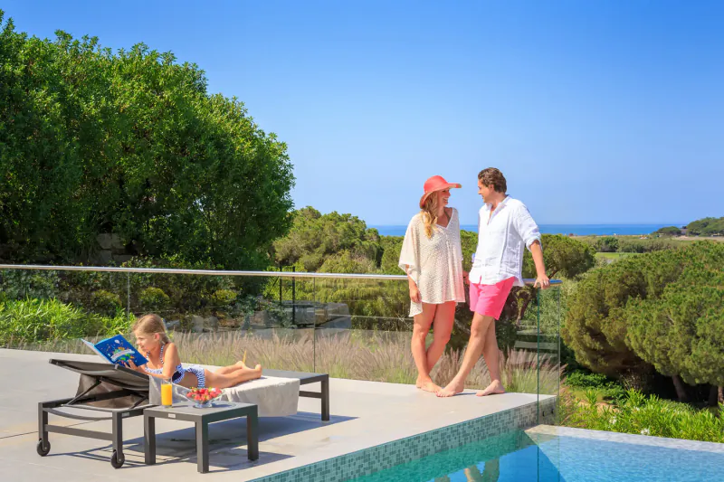 Family at Vale Do Lobo Resort: girl reading on lounge chair by pool, parents in sunhat and shorts standing on deck overlooking ocean.
