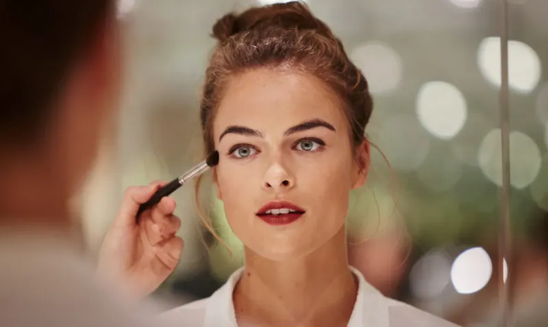Makeup artist applying eyeshadow to woman's eye in salon mirror reflection