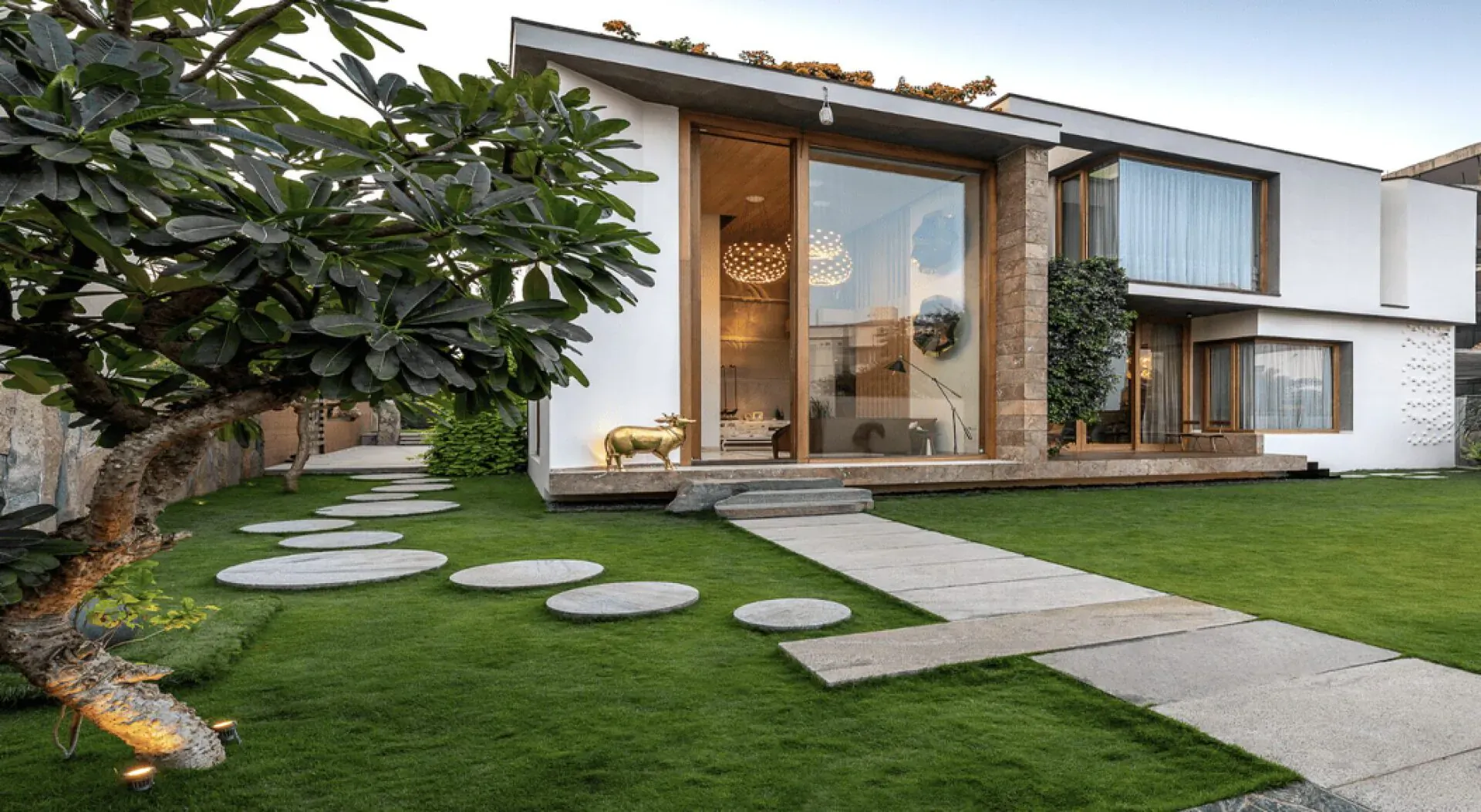 Modern white house with large glass windows, green roof, lush lawn, stepping stone path, and trees under blue sky