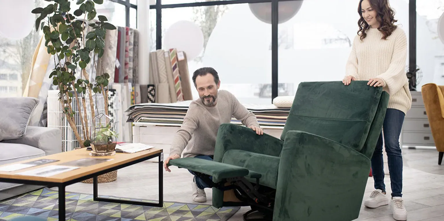 Man and woman lifting green velvet recliner chair in modern showroom with plants and furniture.