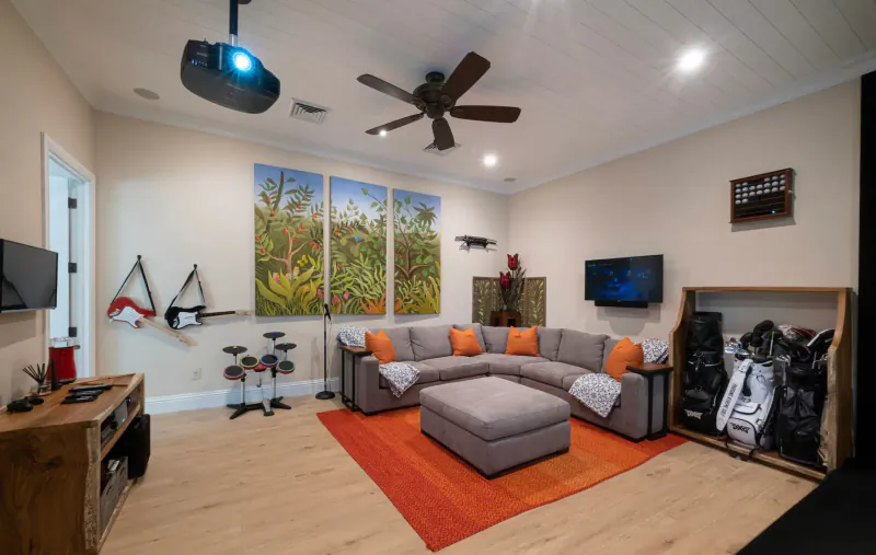 Spacious luxury villa living room with gray L-shaped sofa, orange rug, projector, golf bags, drum kit, and tropical art.