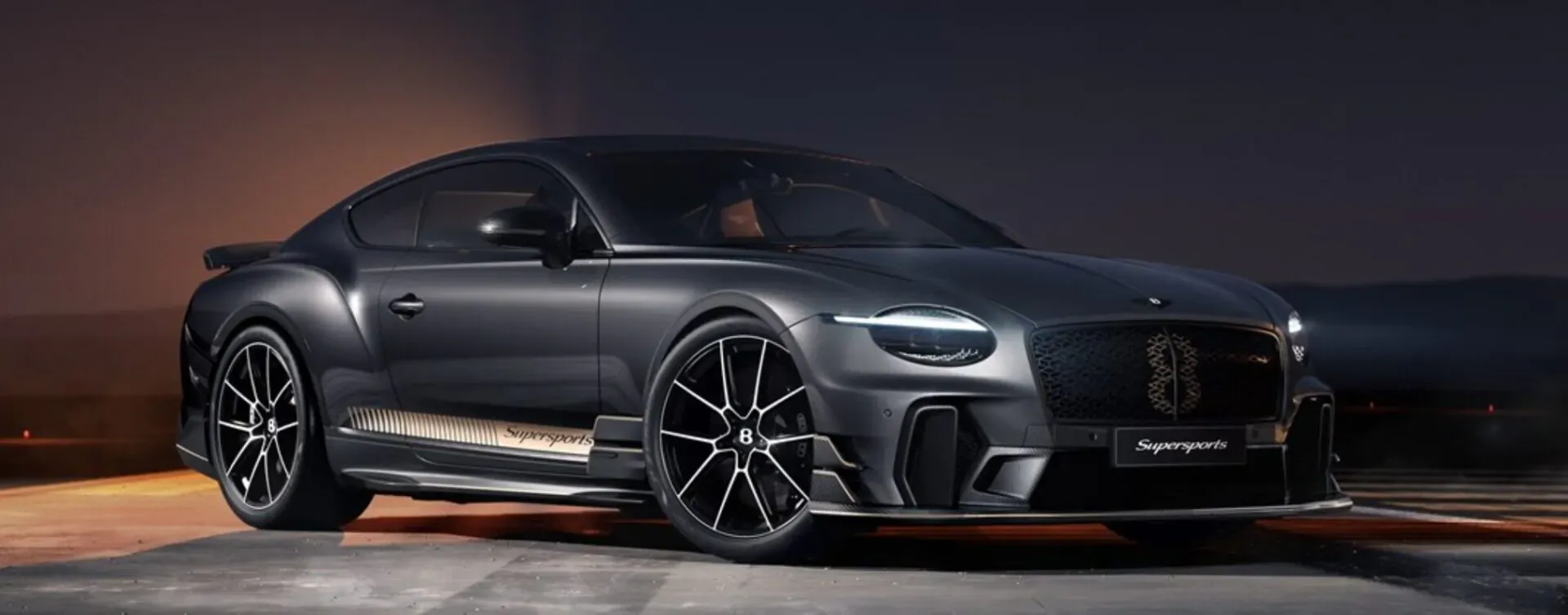 Sleek dark grey Bentley Supersports coupe with large black wheels, parked at night with warm lighting glow.