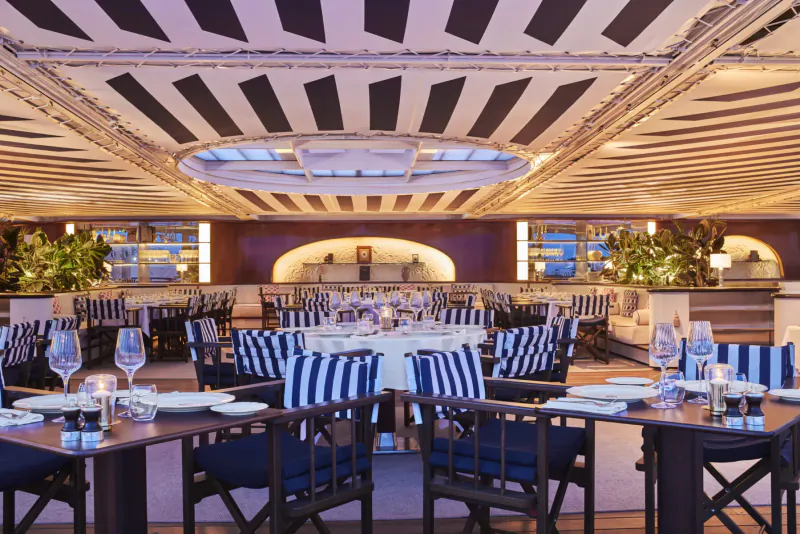 Elegant empty restaurant with striped ceiling, round skylight, navy chairs, white tables, and warm lighting.
