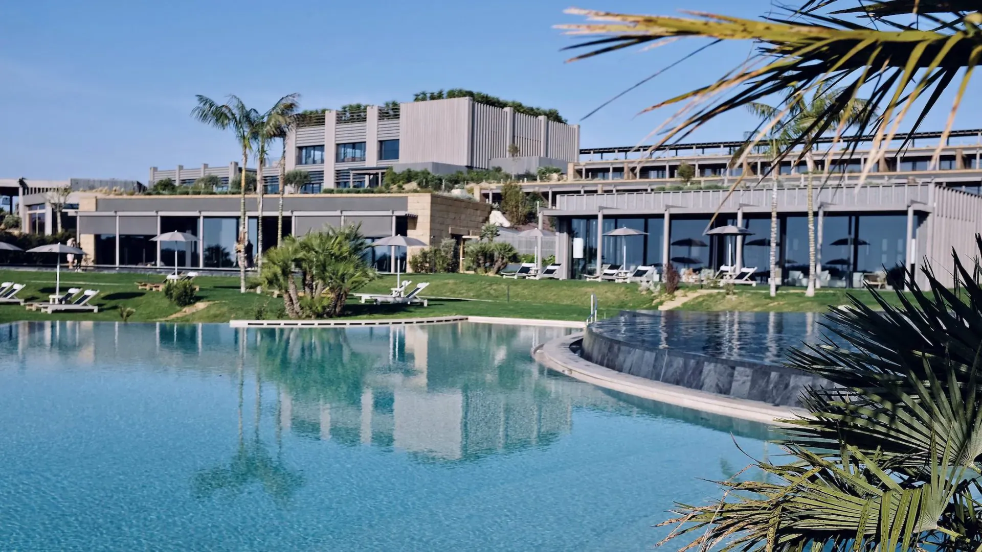 Aerial view of ADLER Spa Resort Sicily: modern terraced buildings, turquoise pool, palm trees, green lawns under blue sky.
