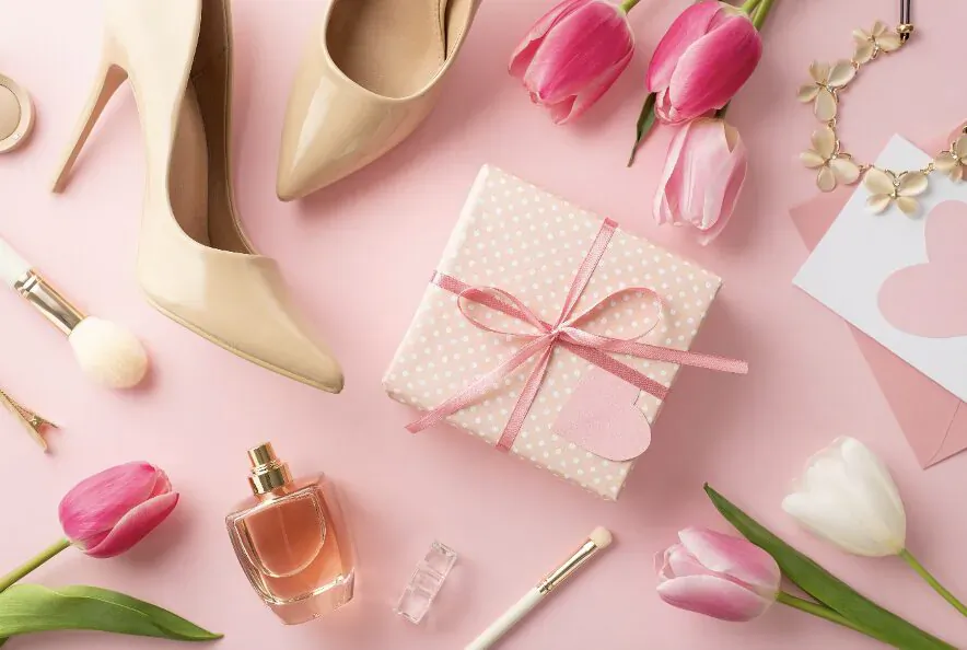 Flat lay on pink background: pink tulips, nude high heels, perfume bottle, gift box with bow and heart card for Valentine's Day