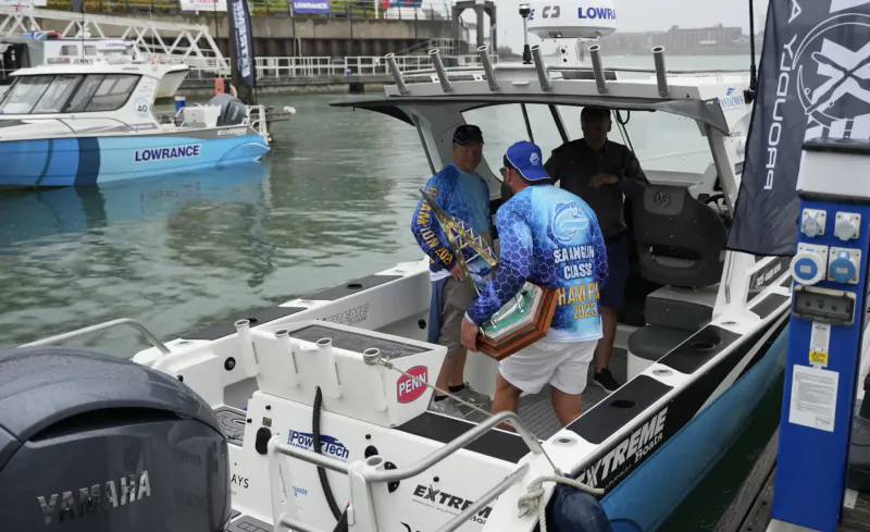 Men in fishing gear on white Quintrex Extreme boat at marina dock, holding green box, other Lowrance boats nearby, End of Summer Boat Show.