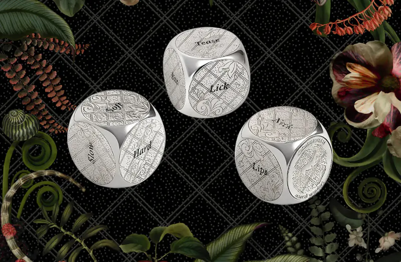 Three engraved silver dice reading 'Lust', 'Gluttony', 'Avarice' on black checkered background with lush Jardin d’Éden plants.