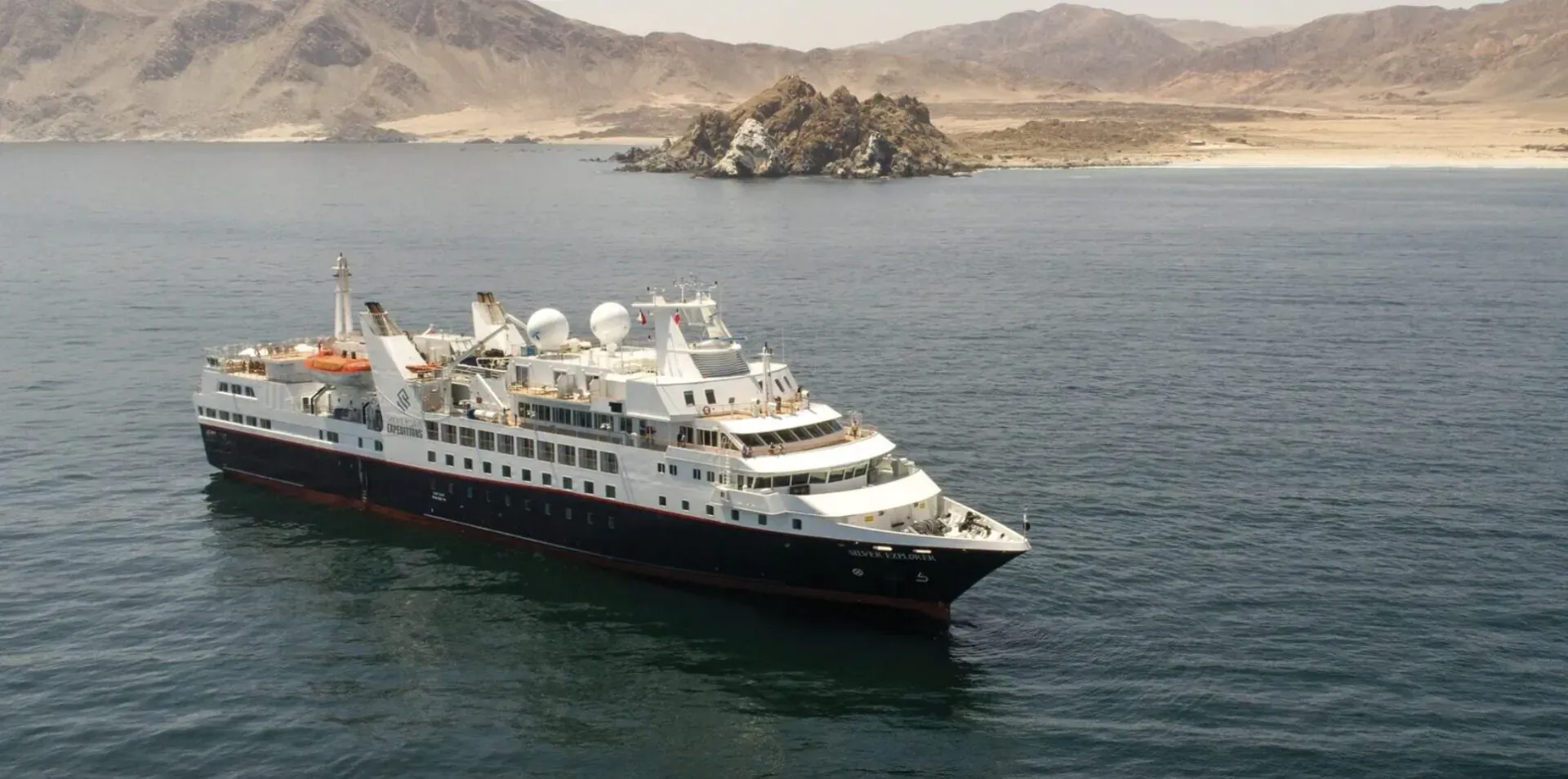 Luxurious white cruise ship with black hull sailing in ocean near rocky desert mountains and beach