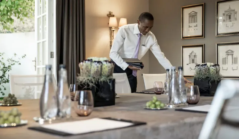 Waiter in white shirt and purple tie arranges glasses, grapes, and potted plants on elegant dining table in Four Seasons Westcliff room.