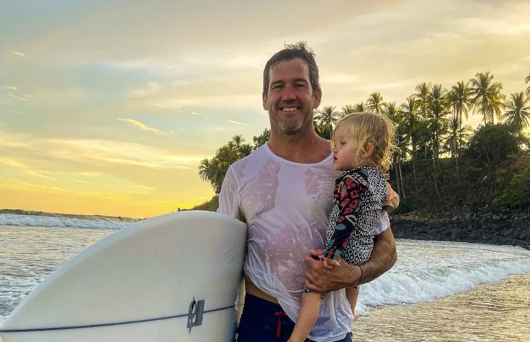 Frank Scotti smiling with toddler daughter on tropical beach at sunset, holding white surfboard, wet shirt, palm trees and waves behind