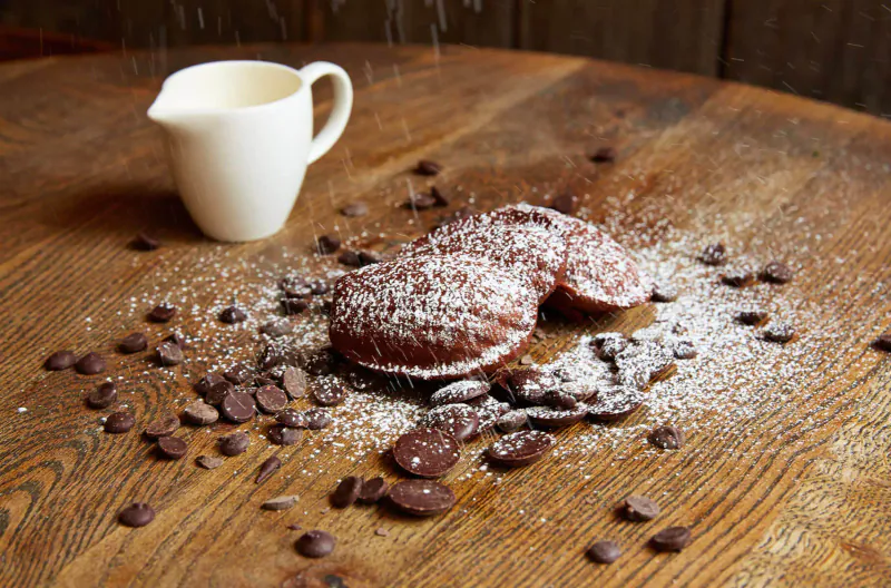Powdered chocolate donuts with scattered chocolate chips on wooden table, white milk jug pouring powdered sugar nearby.