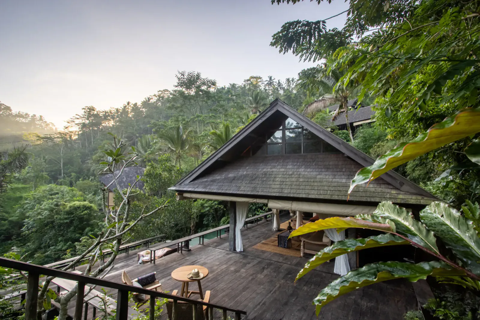 Wooden wellness bungalow with deck, table, and chairs overlooking lush misty Bali jungle at sunrise