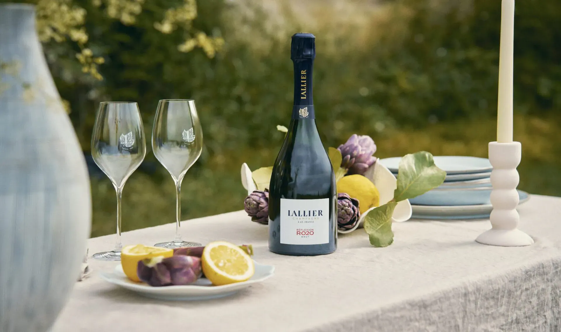 Champagne Lallier bottle with two glasses, lemon slices, artichokes on table outdoors for National Champagne Day.