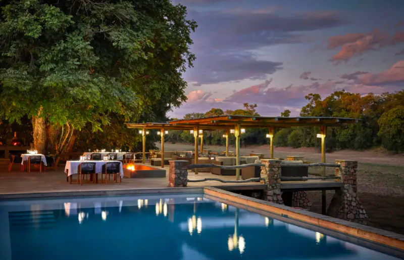 Twilight view of Mfuwe Lodge: infinity pool, outdoor dining under pergola, fire pit, trees, and bushveld riverbank.