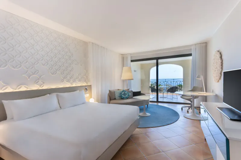 Elegant Hilton Malta hotel room with white king bed, lounge area, desk, TV, and sea view balcony.