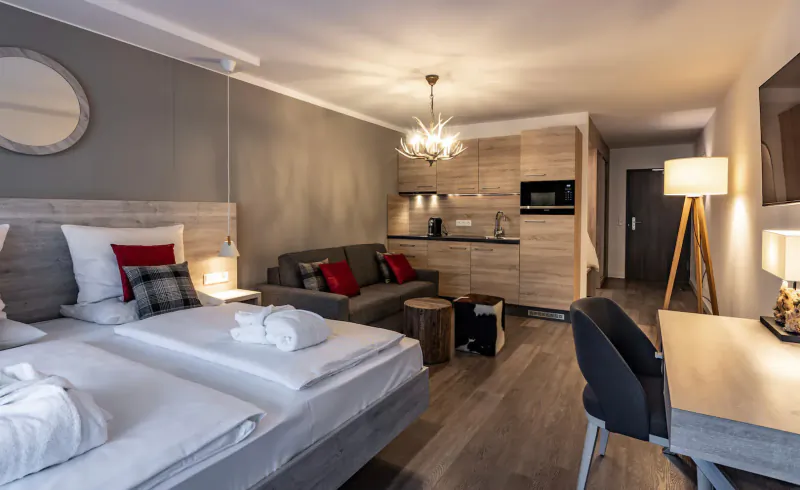 Cozy modern suite at Karma Bavaria with twin beds, red sofa, kitchenette, desk, and warm lighting.