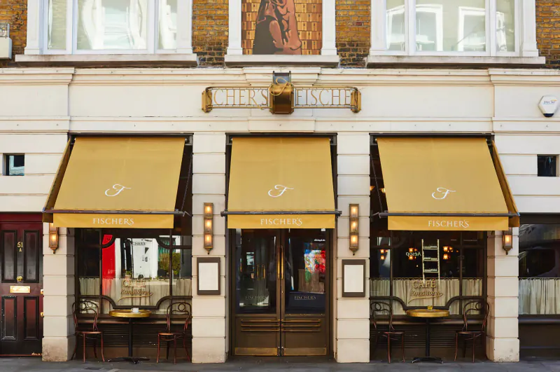 Elegant restaurant facade in Marylebone Village with three gold awnings marked 'F', ornate sign, tables, and lamps.