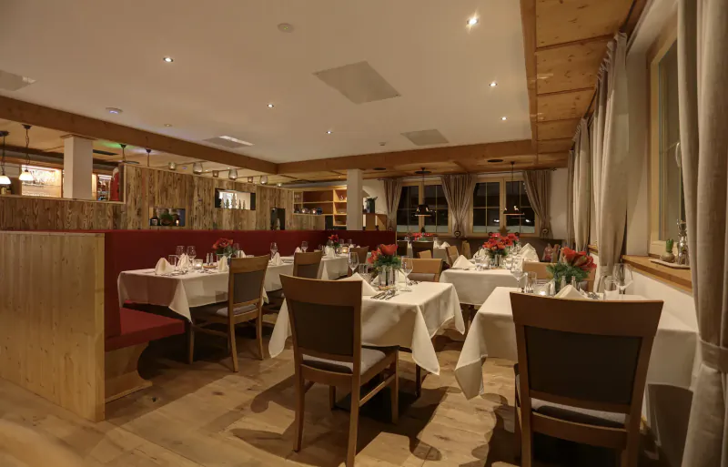 Cozy alpine hotel dining room with wooden beams, red booths, white-clothed tables set with wine glasses and red roses.