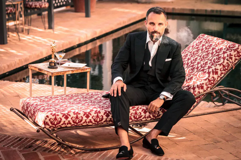 Man with goatee in black suit and vest sits on red chaise lounge by pool, Savile Row Oscar on table, Marbella setting