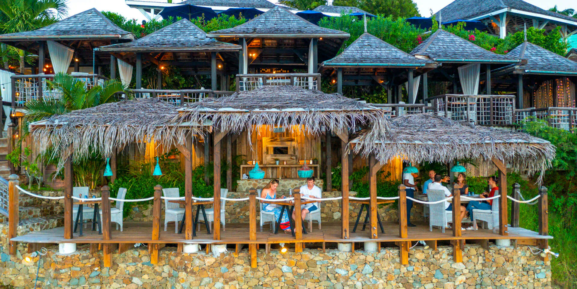 Sheer Rocks in Antigua: thatched beachfront bar and deck with guests dining, overlooking turquoise sea.
