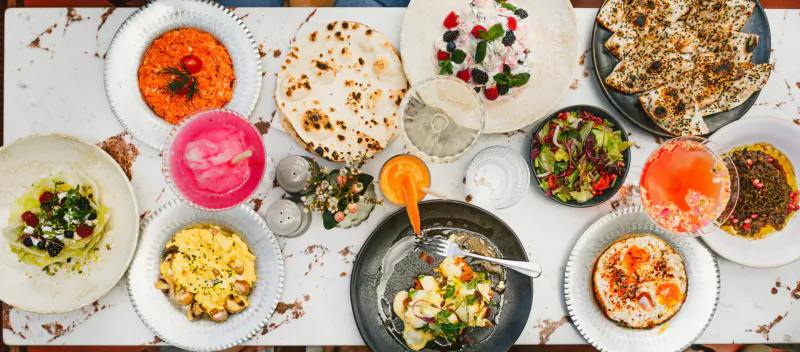 Overhead view of Villa Mamas spread: colorful Middle Eastern dishes including naan, hummus, salads, soups on rustic table.