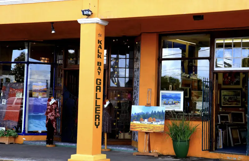 Kalk Bay Gallery exterior in yellow building with artworks in windows, plants, and people outside, Cape Town.