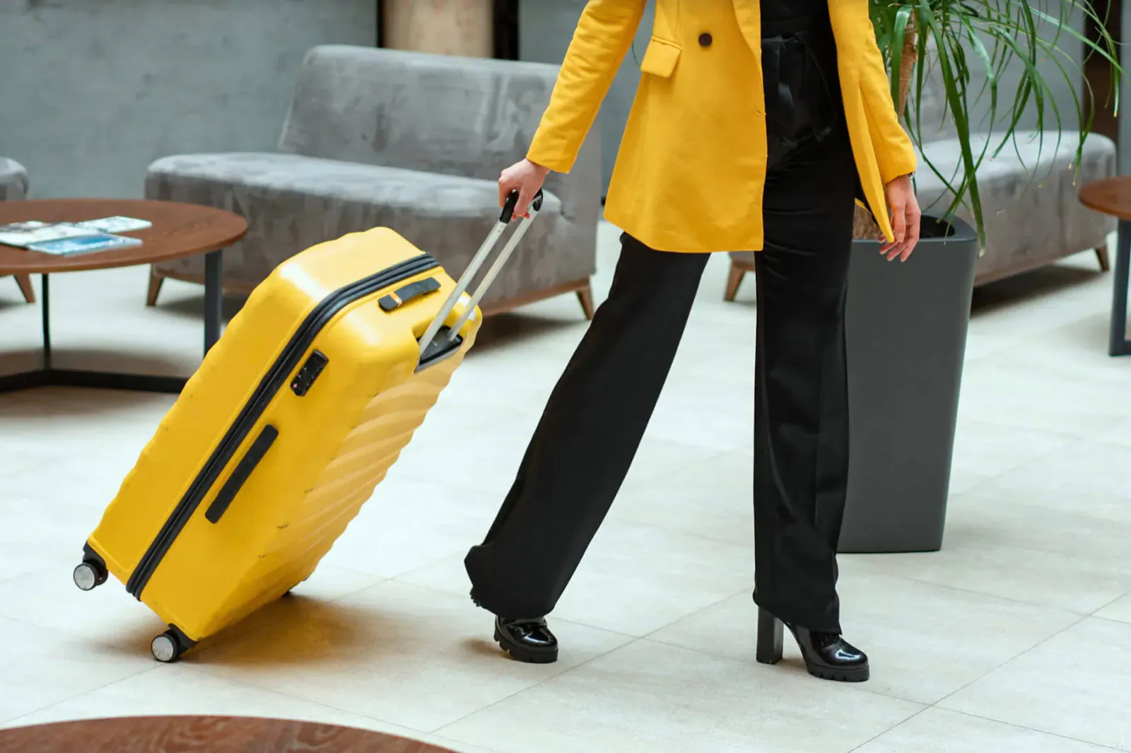 Woman in yellow coat and black pants pulling yellow suitcase in modern hotel lobby with plants and seating.