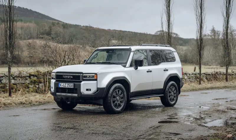 White Toyota Land Cruiser SUV parked on wet rural road with hills, trees, and stone wall backdrop