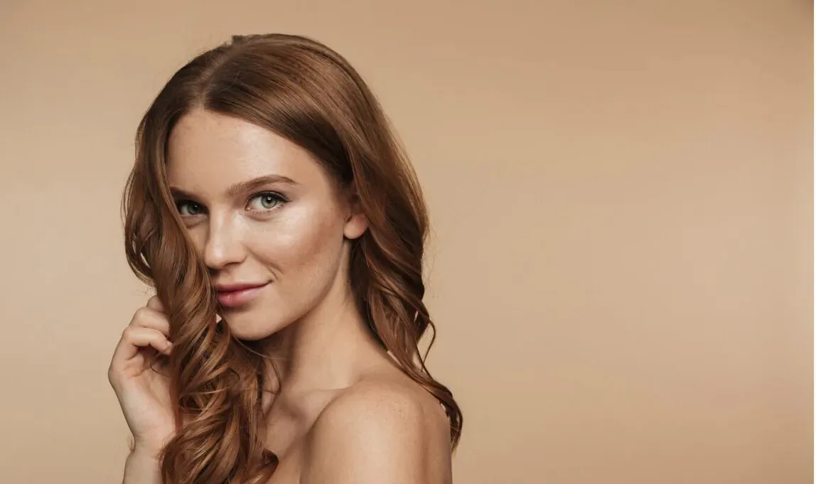 Close-up portrait of smiling woman with long wavy red hair touching her locks against beige background