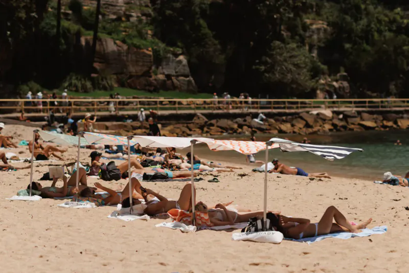 Crowded beach with diverse sunbathers lounging under striped umbrellas and towels, rocky cliffs and ocean backdrop.
