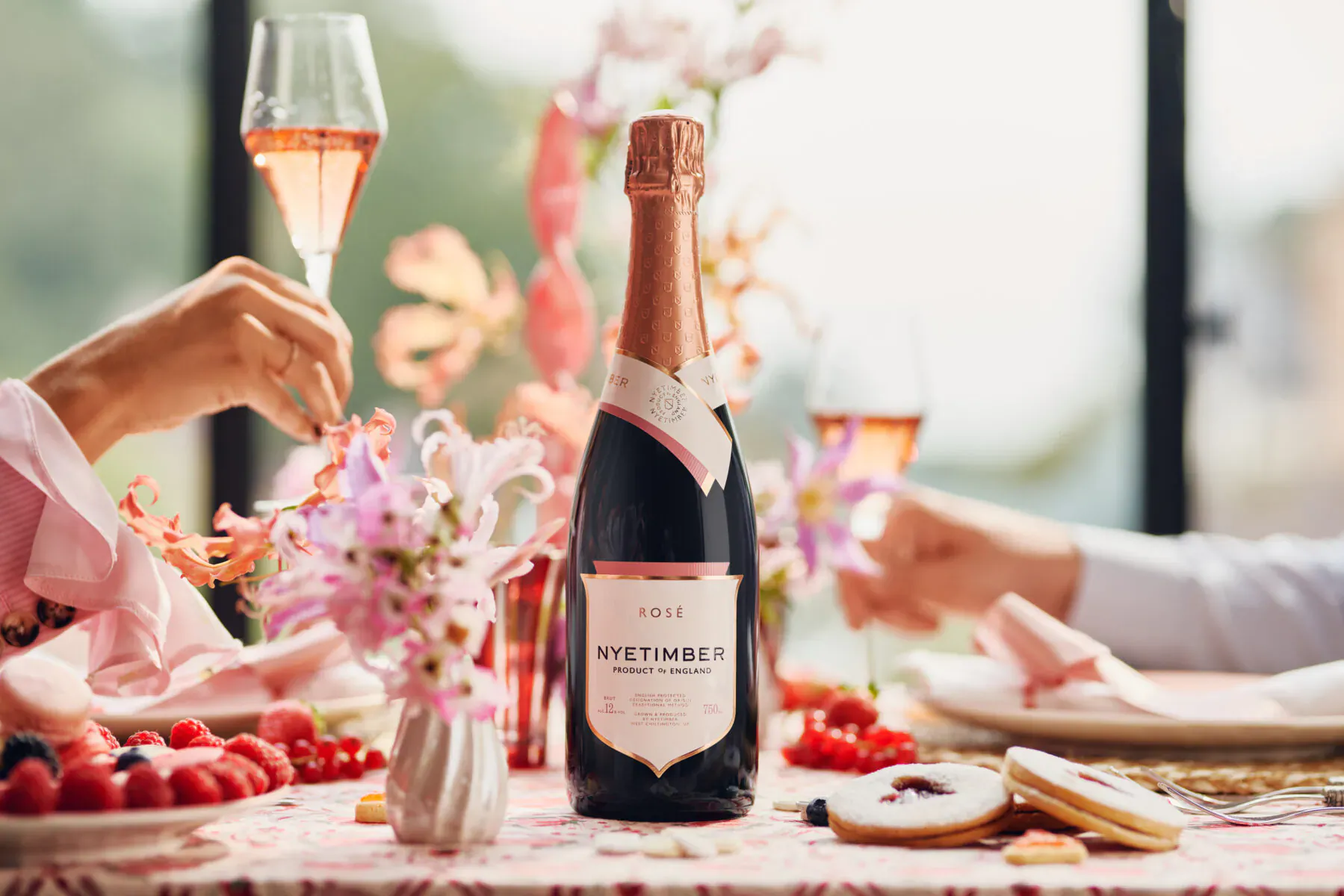 Nyetimber 2018 rosé sparkling wine bottle on table with pink flowers, flutes of rosé champagne, hands toasting, strawberries, for Mother's Day celebration