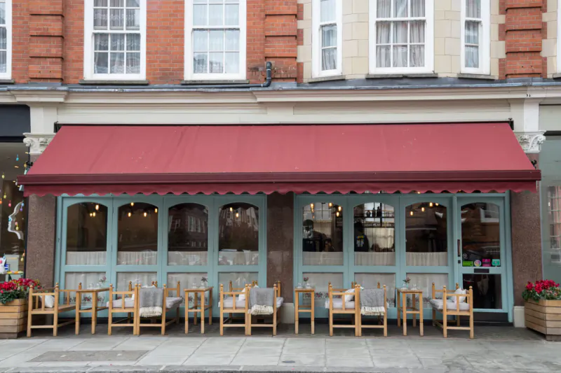 Villa Mamas London café exterior with red awning, teal doors, wooden chairs, and potted red flowers on sidewalk.
