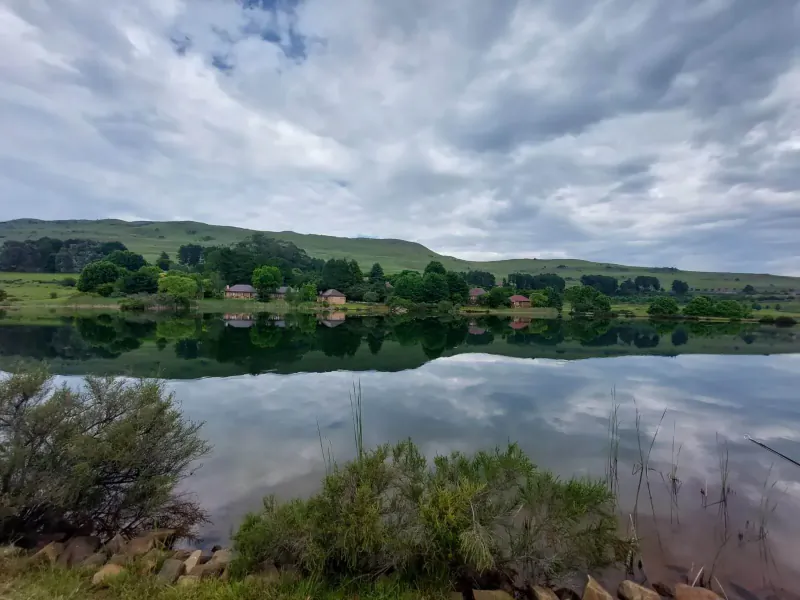 Walkersons Hotel & Spa at Dullstroom: houses reflected in calm lake with green hills under cloudy sky