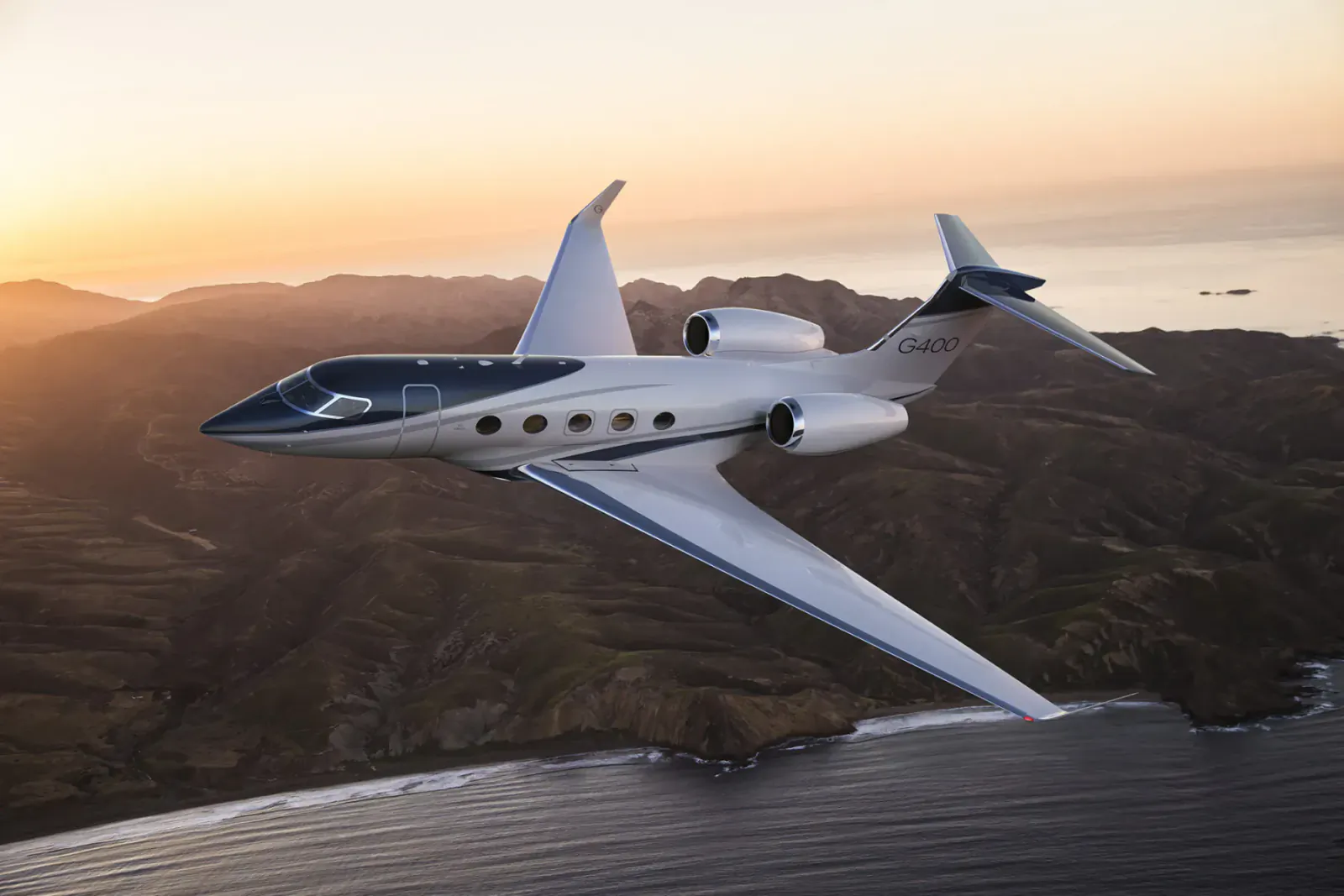 White Gulfstream private jet flying over coastal mountains at sunset with ocean below.