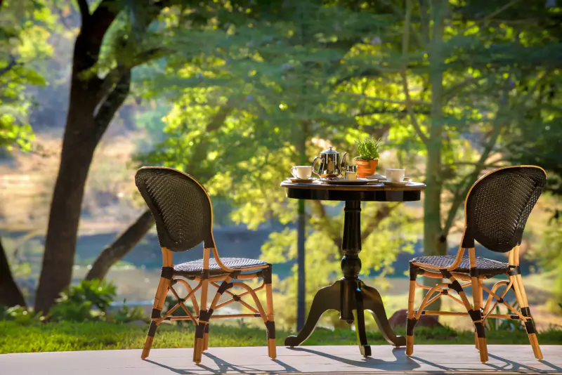 Two wicker chairs at a small round table with teacups and flowers, on a terrace overlooking lush green trees and river at a luxury hotel.
