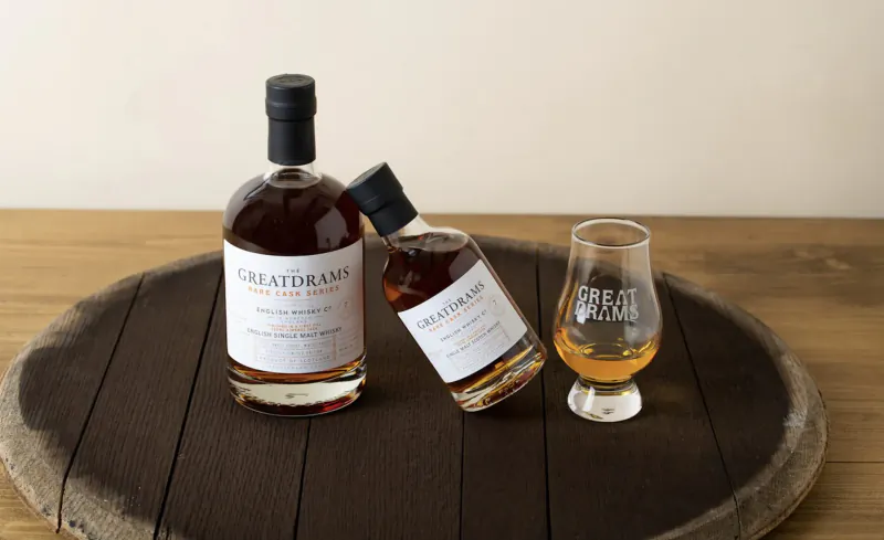 GreatDrams whisky bottles, one tilted with whisky in Glencairn glass, on wooden barrel slice.