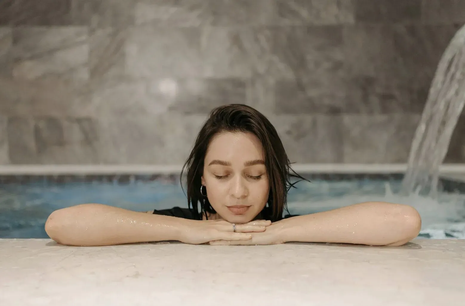 Woman with wet hair relaxes eyes closed, arms folded on pool edge with cascading waterfall behind, tiled spa setting