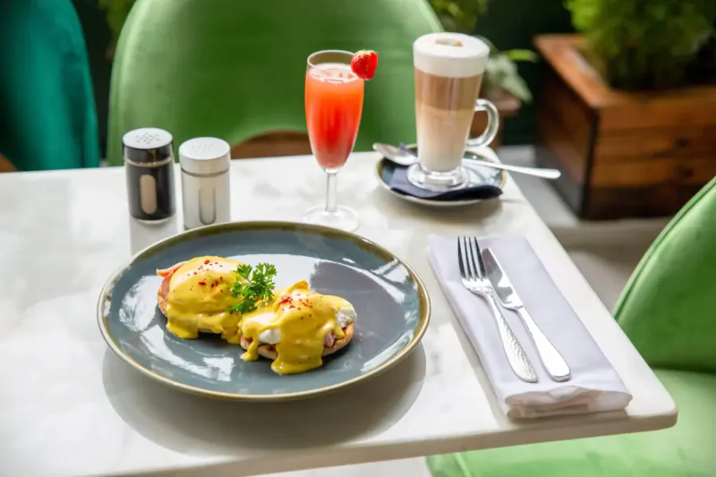 Eggs Benedict with hollandaise on plate, cappuccino, pink juice on marble table at stylish hotel café