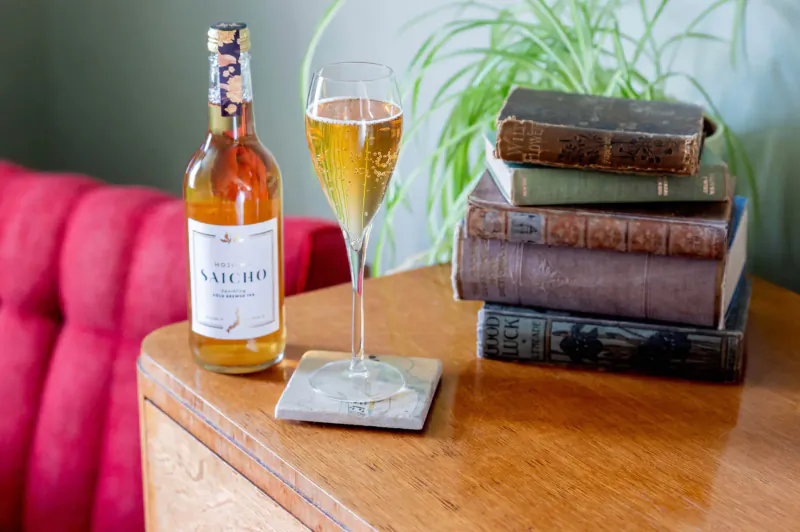 Saicho sparkling tea bottle and poured glass on wooden table with stacked vintage books, green plant, and red chair