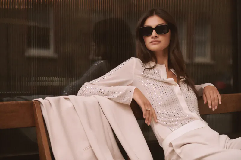 Woman in sheer long-sleeve white blouse and pants sits casually on wooden bench by window in London's St. Christopher's Place gallery.