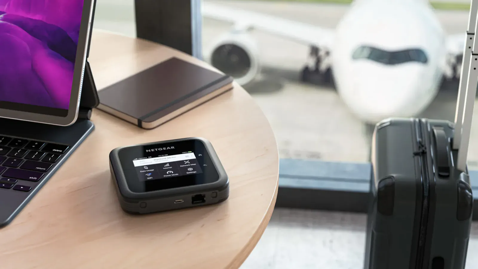 NETGEAR M6 Pro mobile router, purple laptop, notebook, and suitcase on table at airport gate with airplane outside.