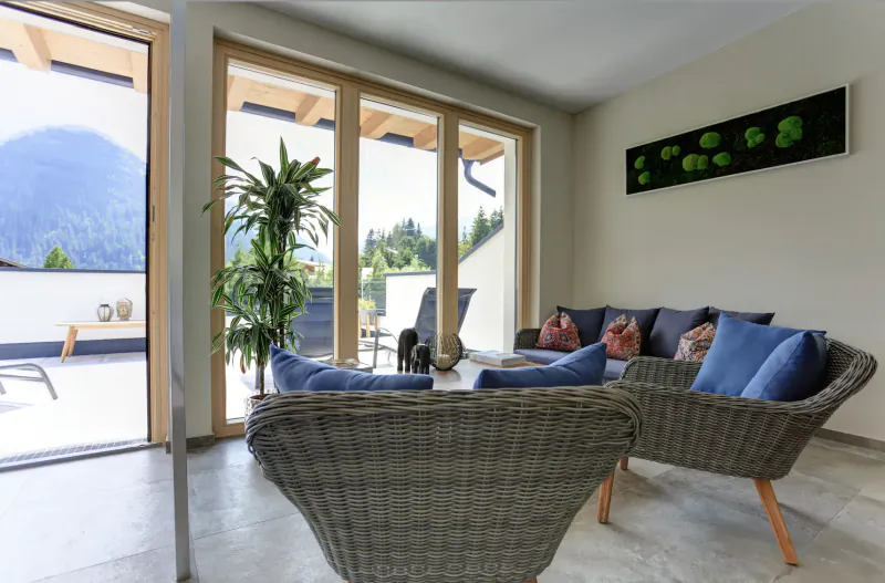 Bright hotel living room with wicker chairs, blue pillows, plants, green wall art, and mountain view through open doors.